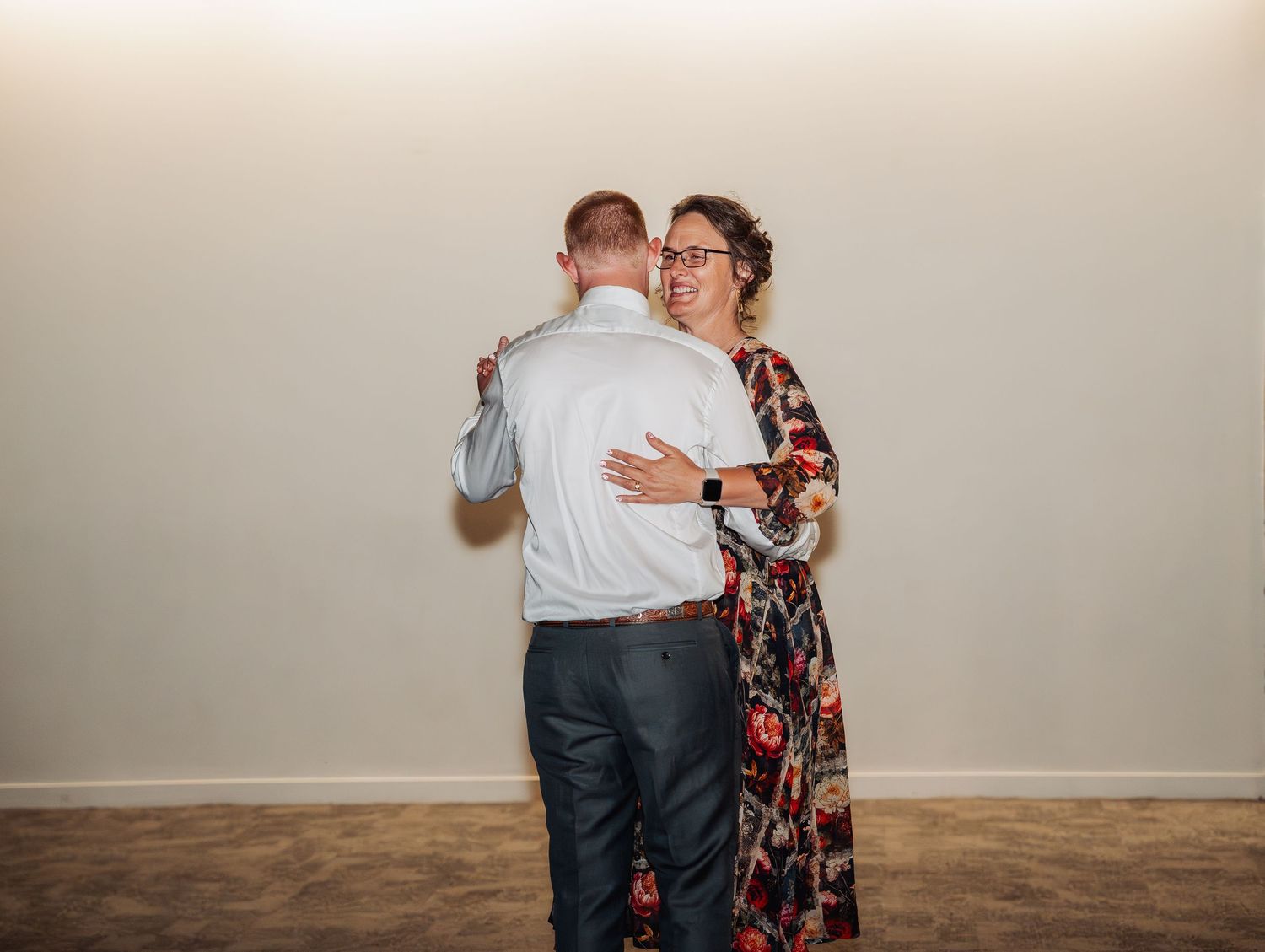 A woman in a floral dress shares a warm dance with a man in a white shirt at an indoor reception.