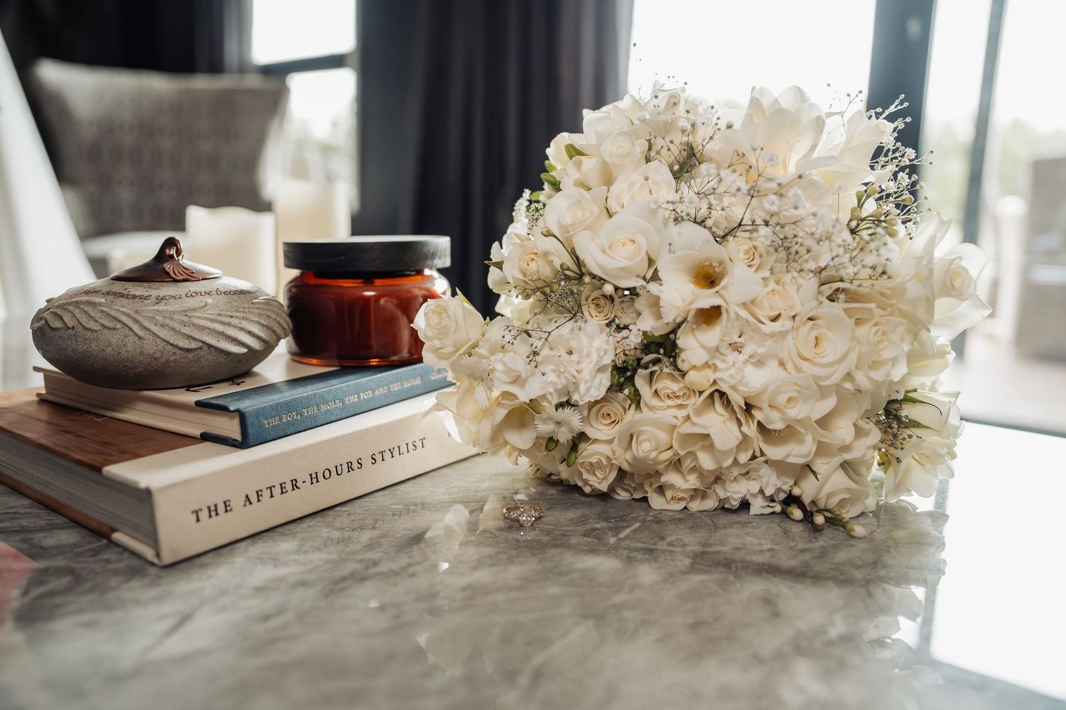 White wedding bouquet with roses and calla lilies rests beside stacked books and candle on marble table.