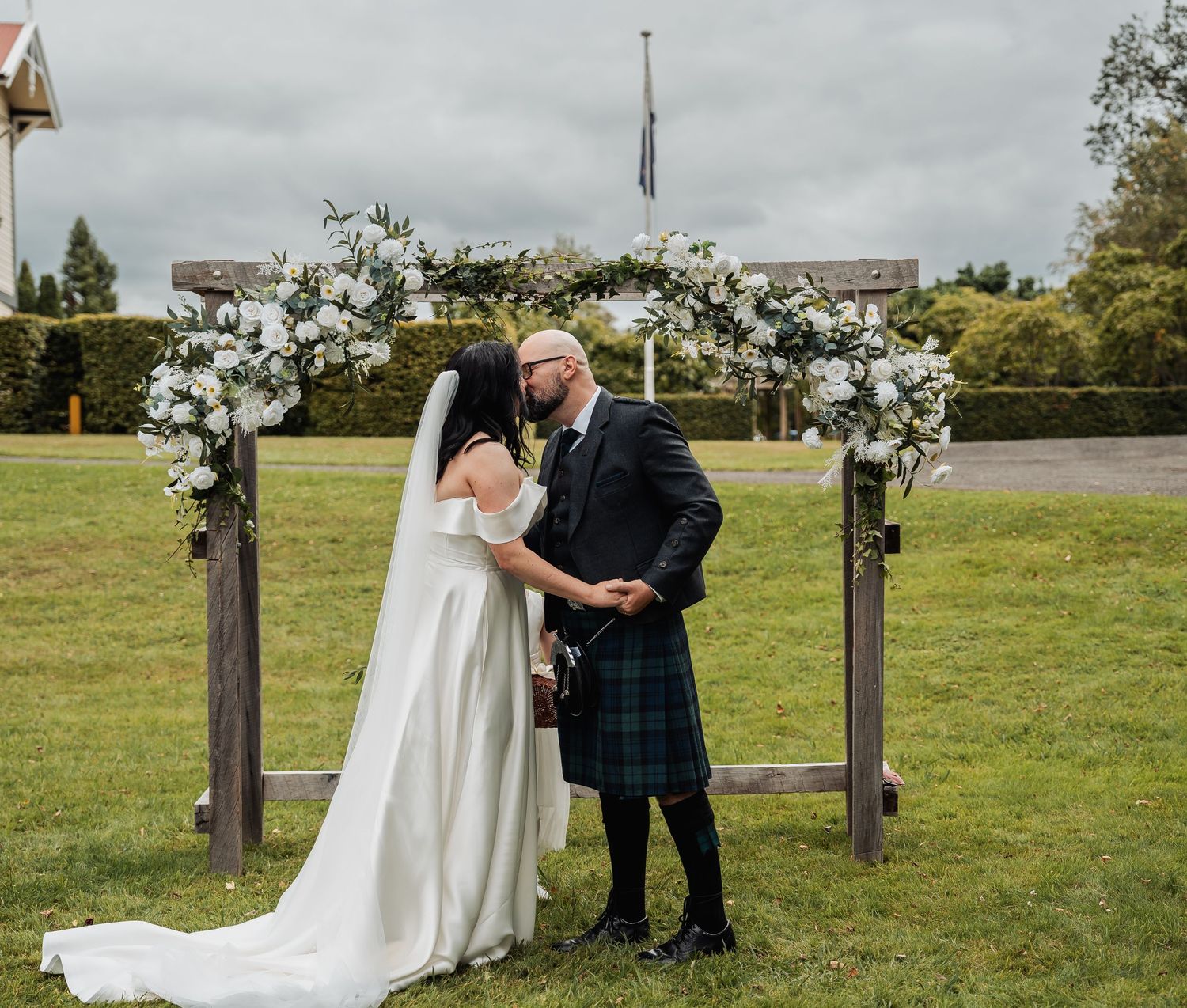 Bride and groom share a kiss under floral arch at outdoor Scottish wedding ceremony, groom wearing tartan kilt.