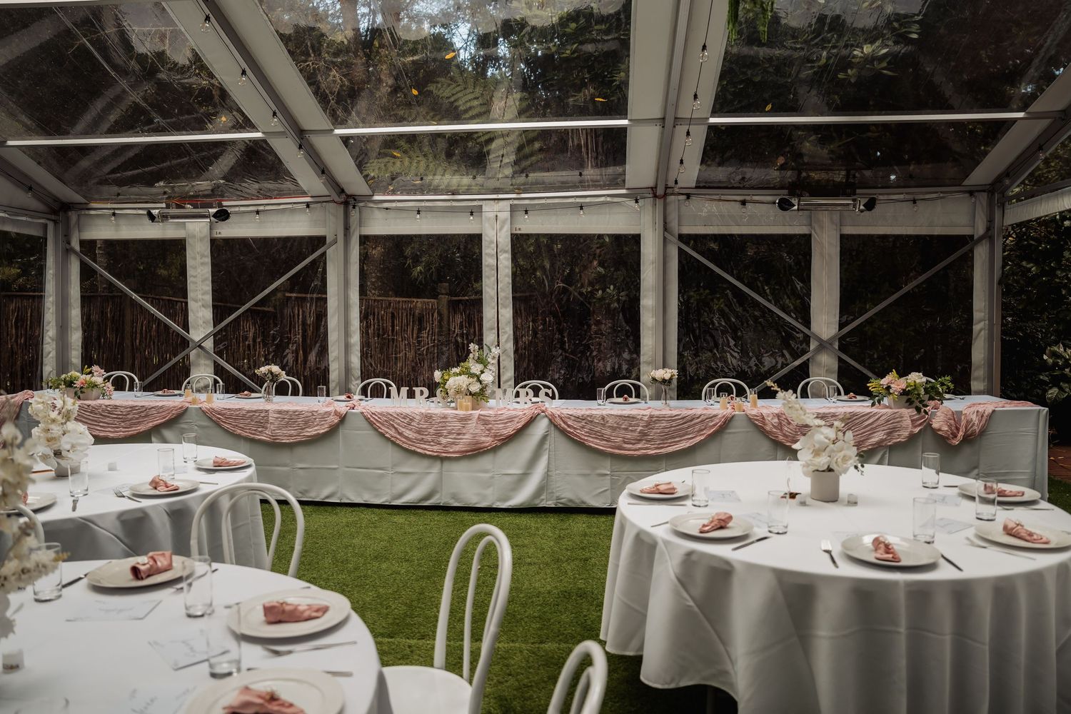 Elegant wedding reception under a clear tent with white round tables, pink napkins, floral centerpieces, and a decorated head table.