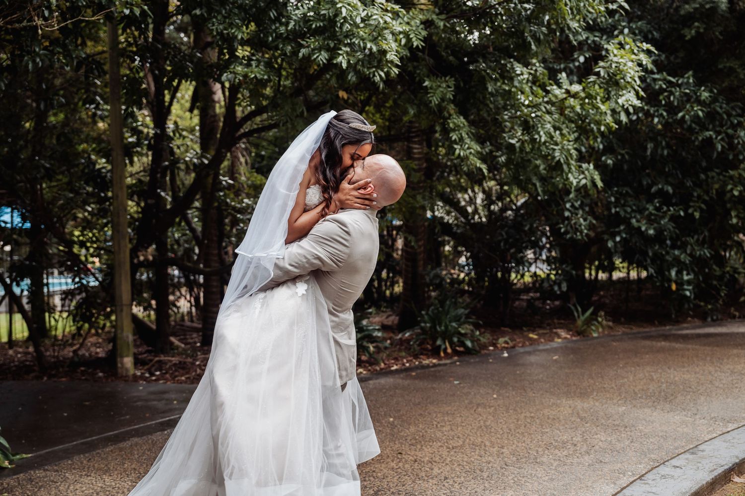 Groom lifts and kisses bride in a romantic outdoor wedding portrait surrounded by lush green trees.