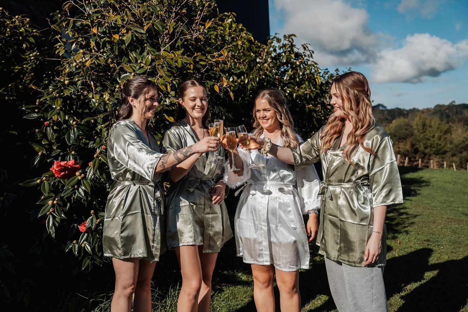 A group in matching silk robes toasts with champagne glasses while standing in a sunny garden setting.