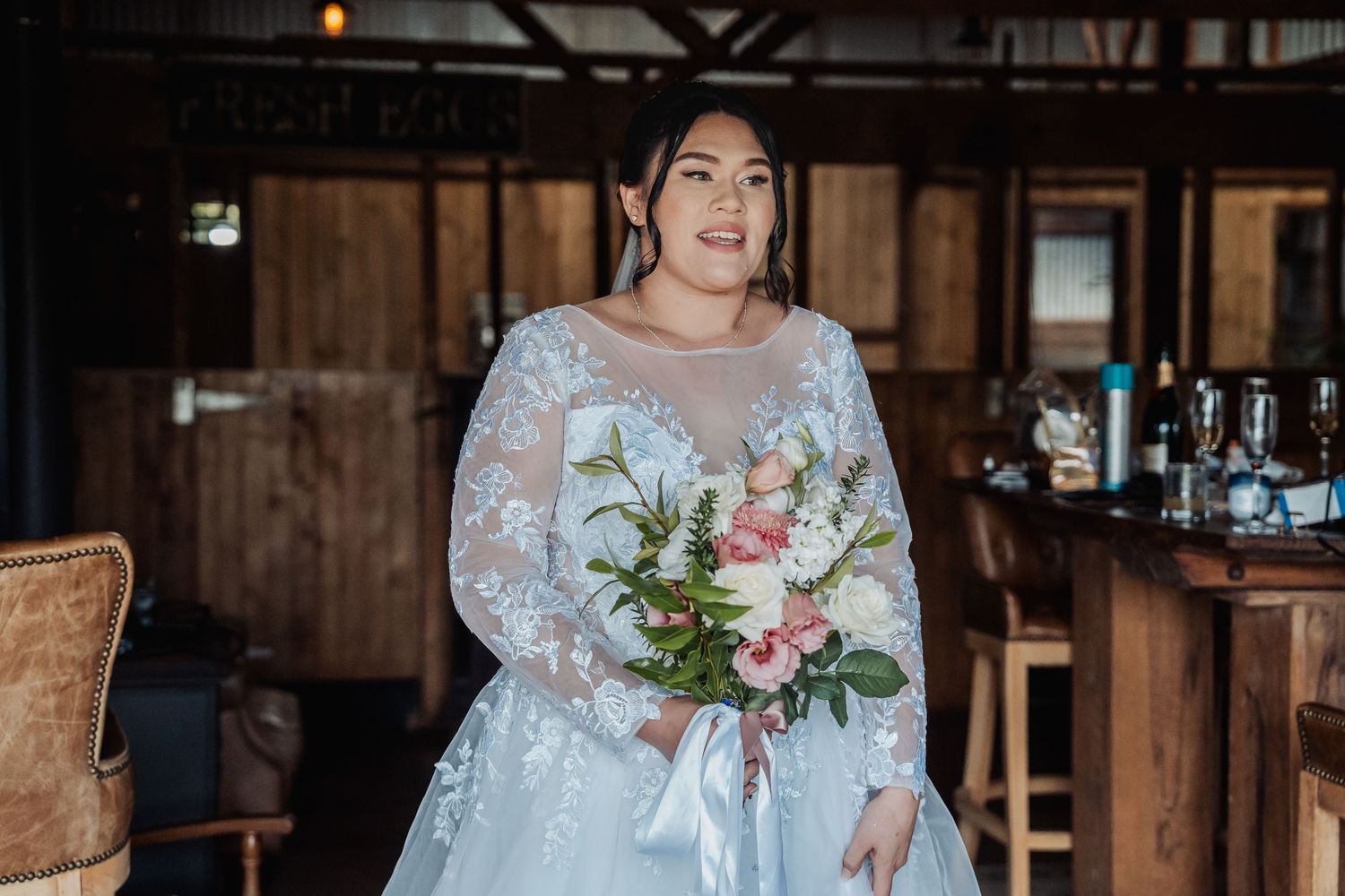 Someone in a detailed lace wedding gown holds a pink and white floral bouquet in a rustic wooden venue.