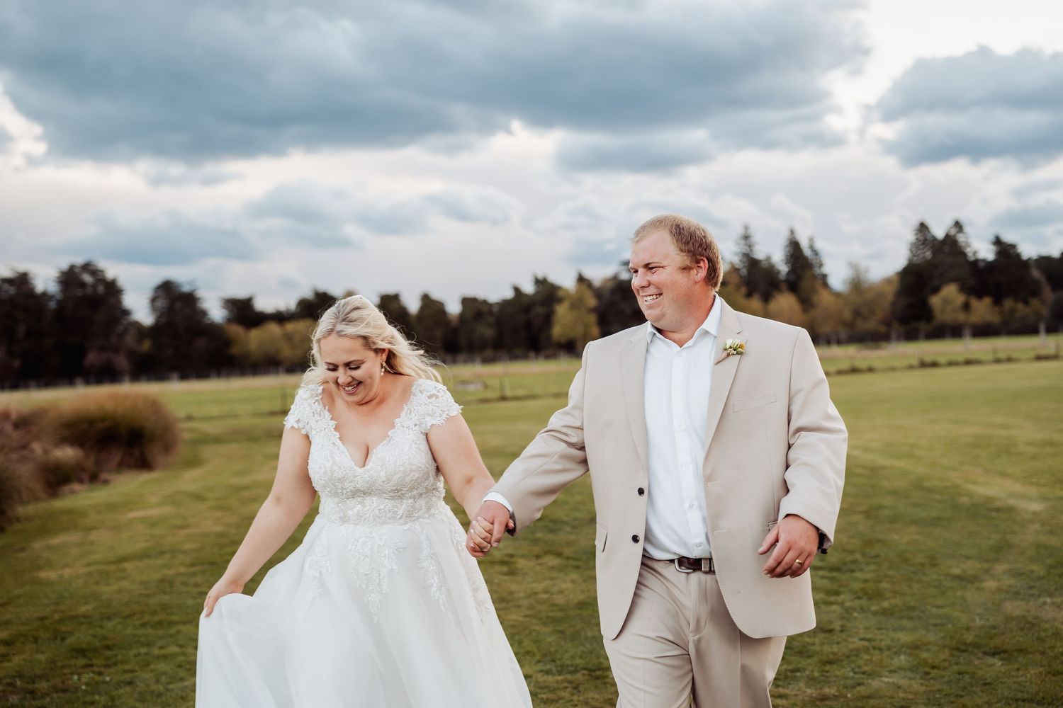 A newlywed couple walks hand in hand across a lush green field under a dramatic cloudy sky after their wedding.