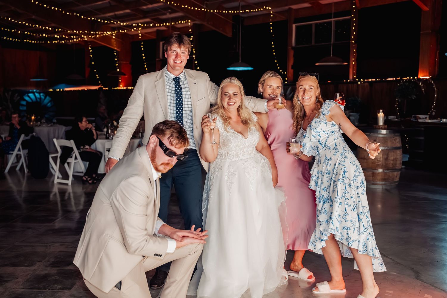 Bride in white gown posing with wedding guests on dance floor at indoor reception with string lights.