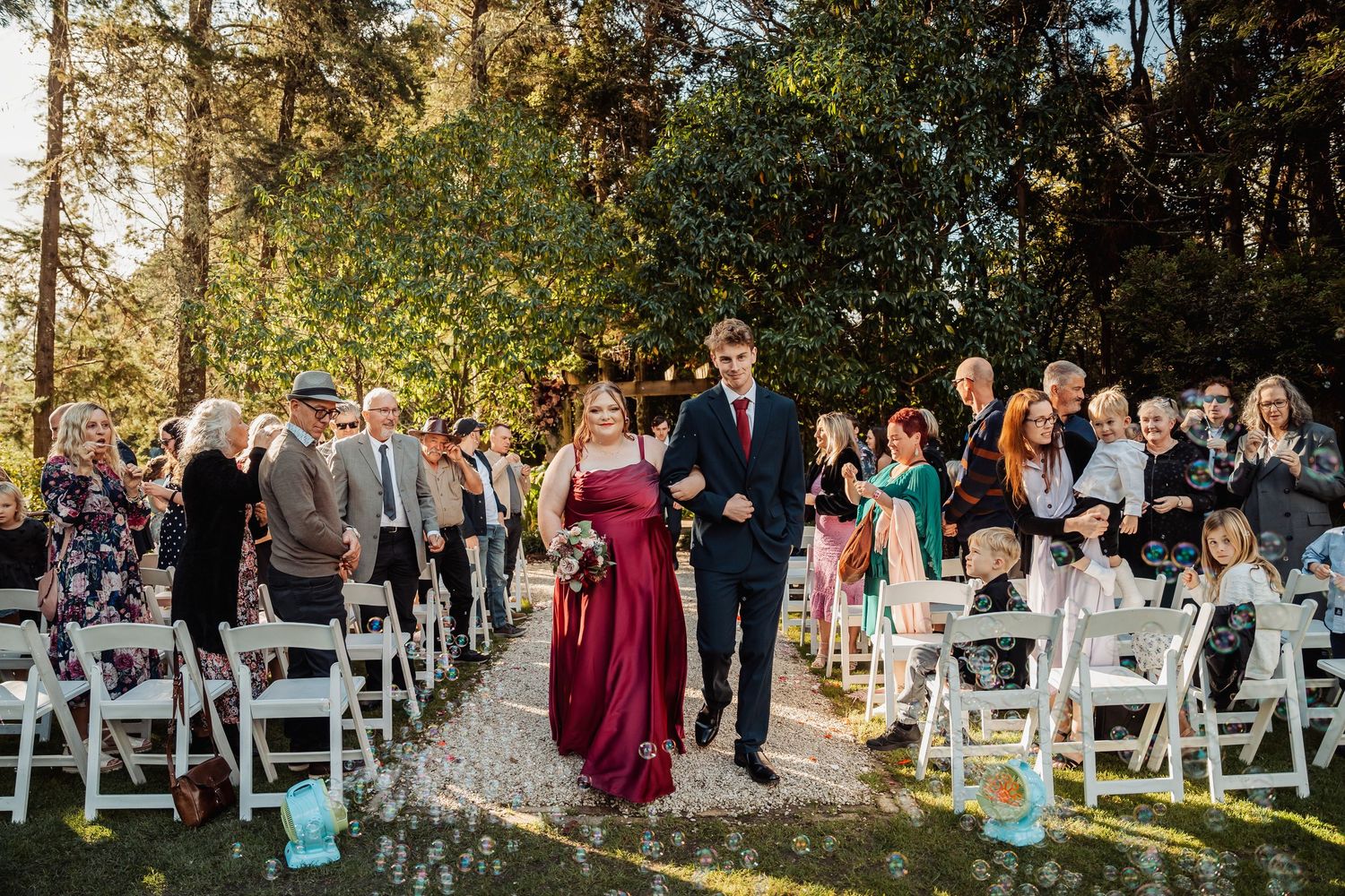 Bridesmaid in burgundy gown walks down outdoor aisle with groomsman past seated wedding guests.