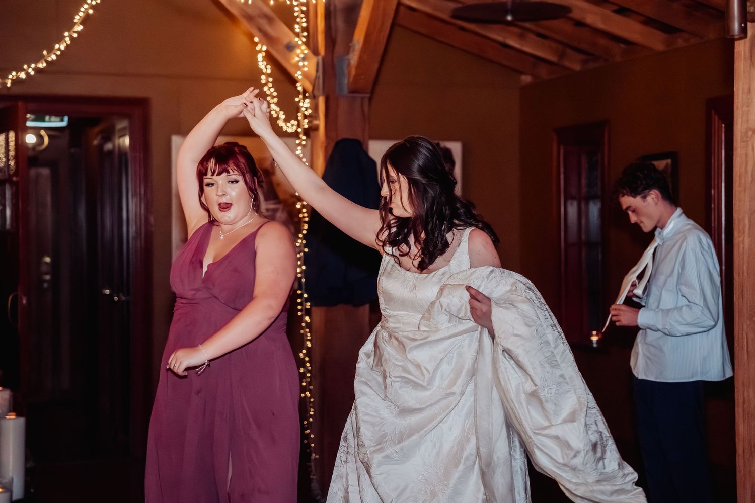 Bride in white gown dancing with bridesmaid in mauve dress at indoor wedding reception.