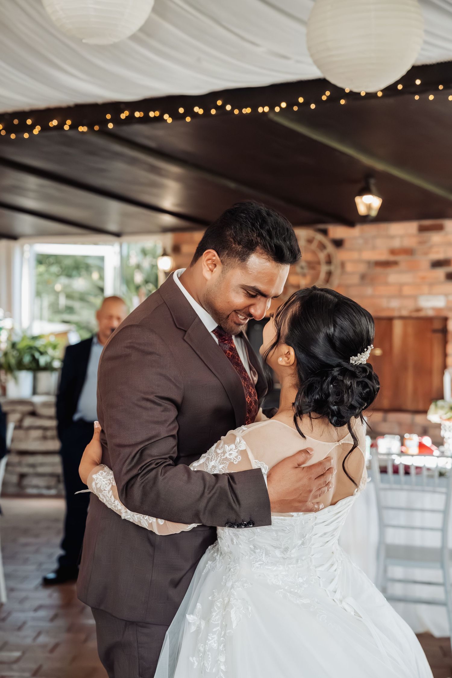 A bride and groom share their first dance, embracing warmly under string lights and paper lanterns at a rustic wedding venue.