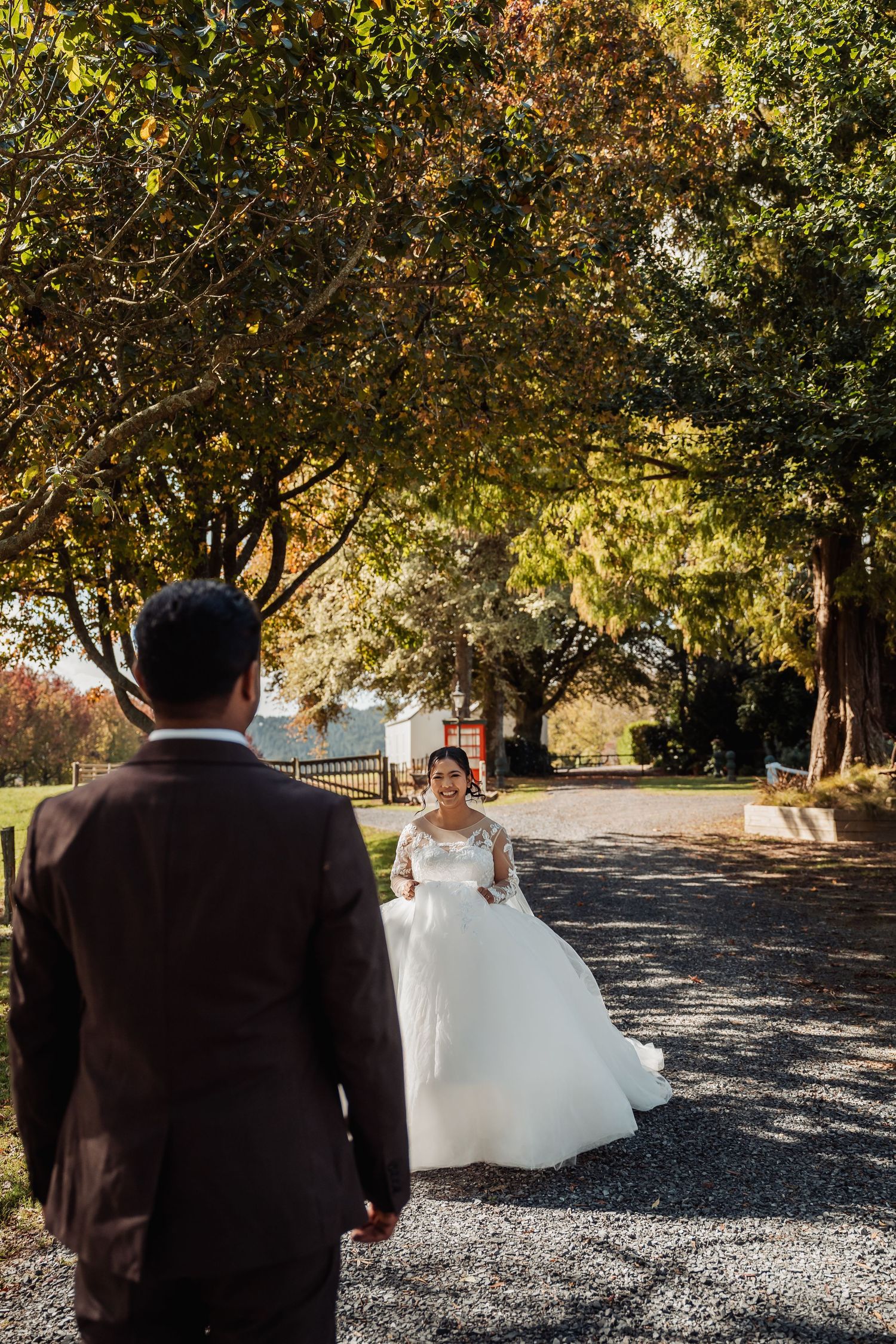 Groom in dark suit watches smiling bride in white lace gown approach on tree-lined gravel path.