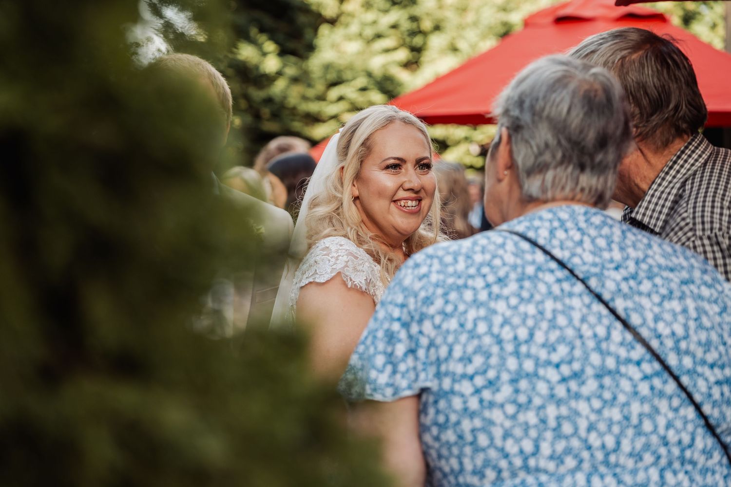 Smiling blonde bride in lace veil mingles with guests near red umbrella at outdoor reception.
