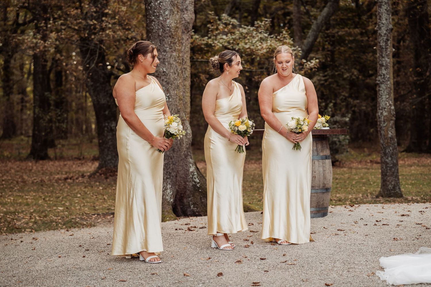 Three bridesmaids in champagne satin one-shoulder gowns holding yellow and white bouquets outdoors among autumn trees.