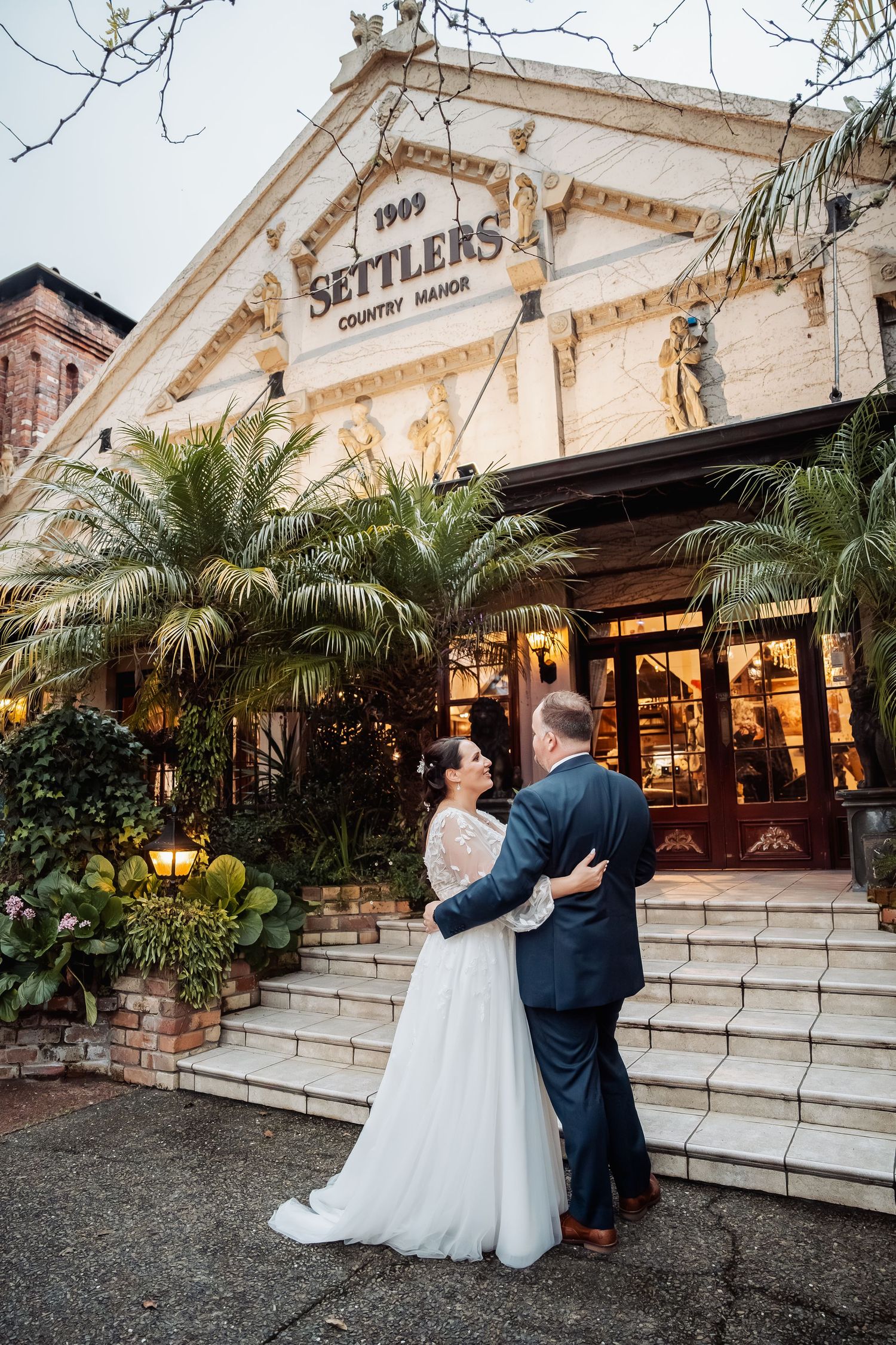 Newlyweds embrace outside The Settlers restaurant with palm trees framing the entrance.