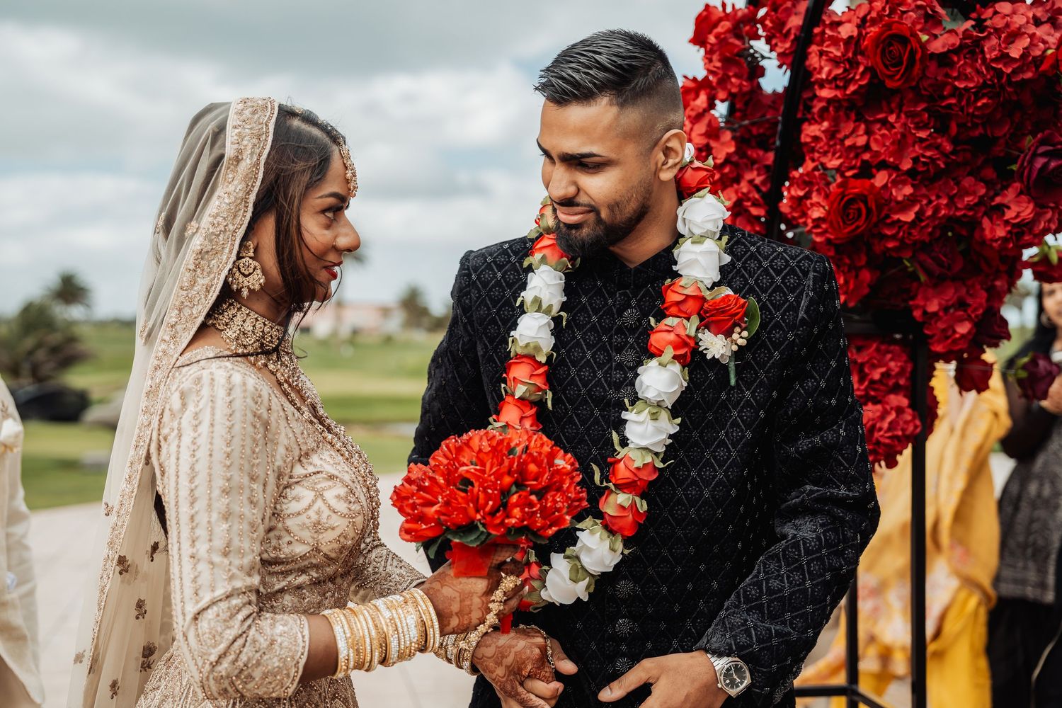 A couple posing together outdoors with vibrant red floral decorations in the background.