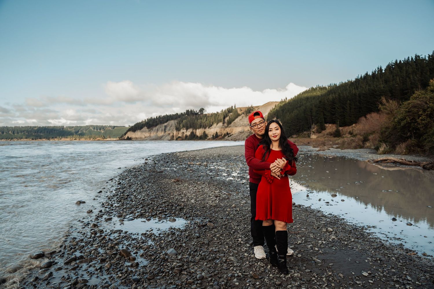 Couple in matching red outfits embracing on a rocky shoreline with forested cliffs and cloudy sky behind them.