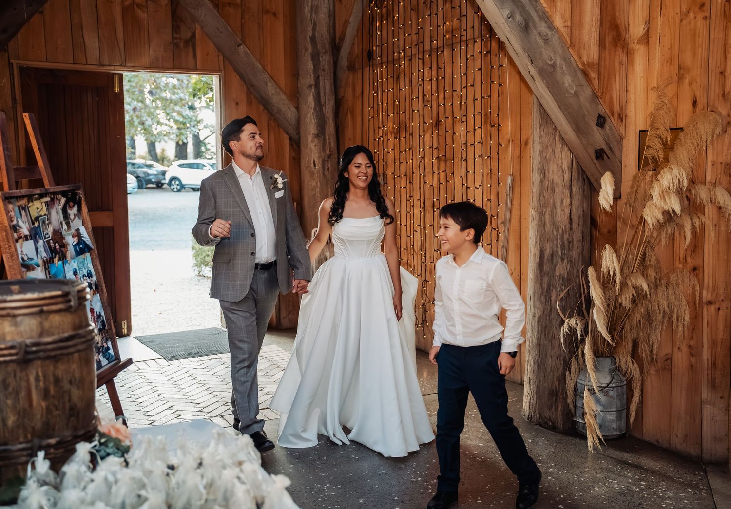 Newlyweds and their son share a joyful moment entering a rustic wooden barn wedding reception venue.