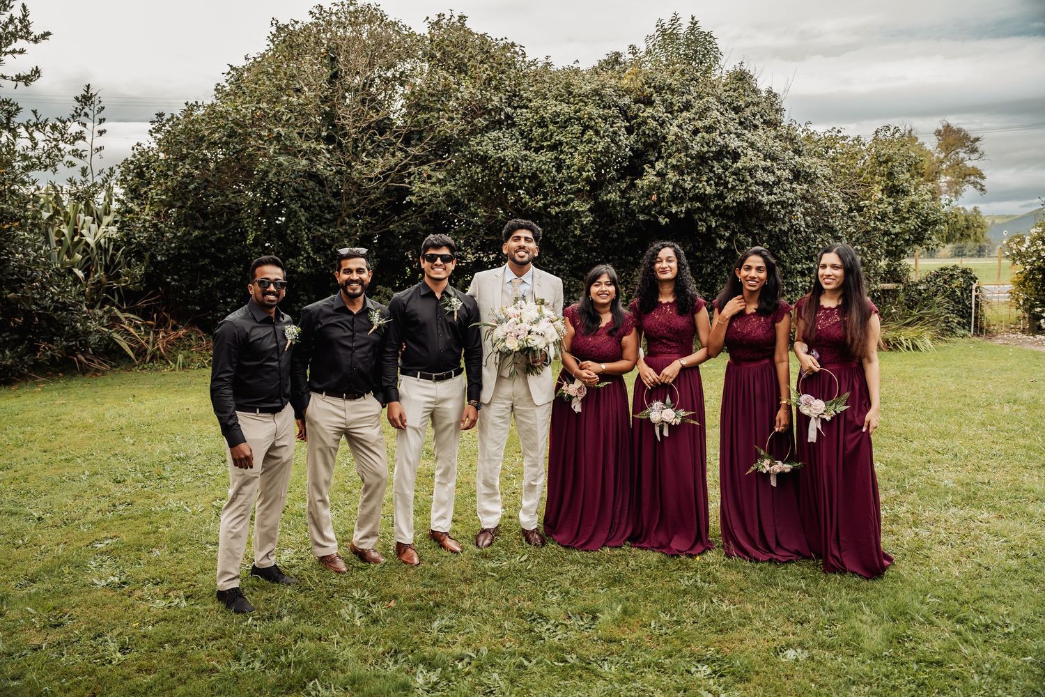 Wedding party posing outdoors on grass, groom in white suit with groomsmen and bridesmaids in burgundy dresses.