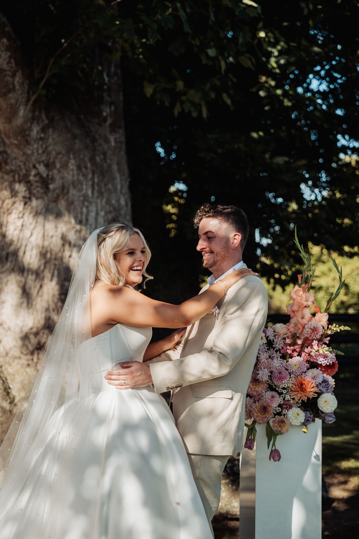 Bride and groom laughing together beside floral arrangements under a large tree at their outdoor wedding.