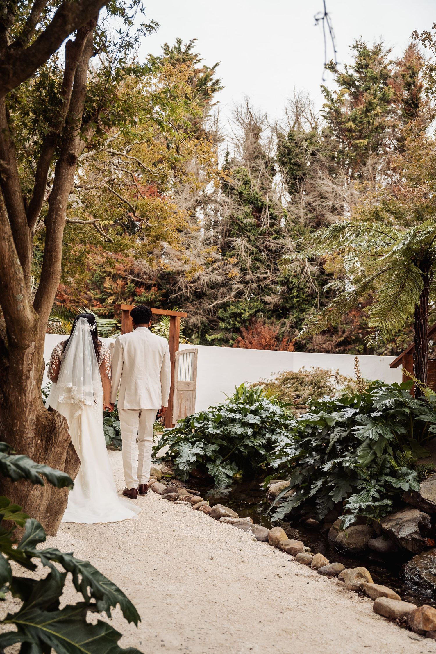 Bride and groom walk hand in hand along garden path surrounded by lush greenery.