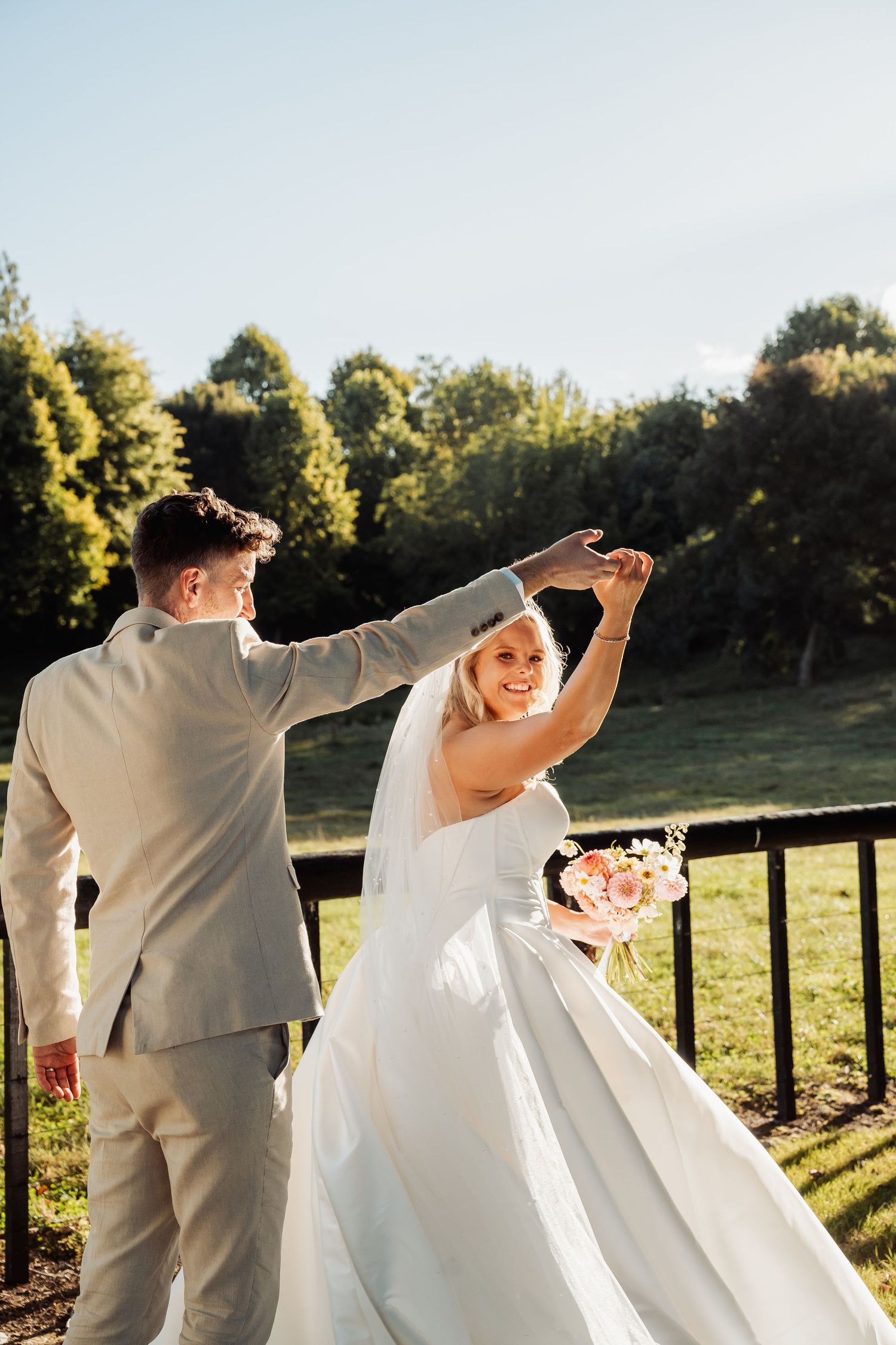 Groom twirls smiling bride in white ballgown outdoors on sunny wedding day.