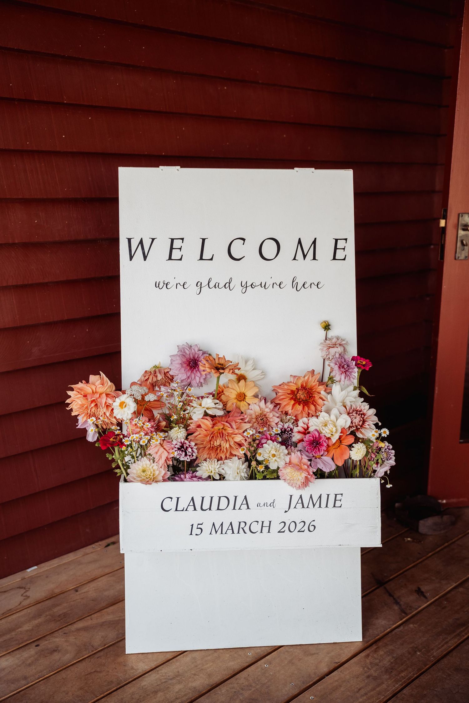 Wedding welcome sign with colorful dahlias for Claudia and Jamie on 15 March 2026 against red barn wall.