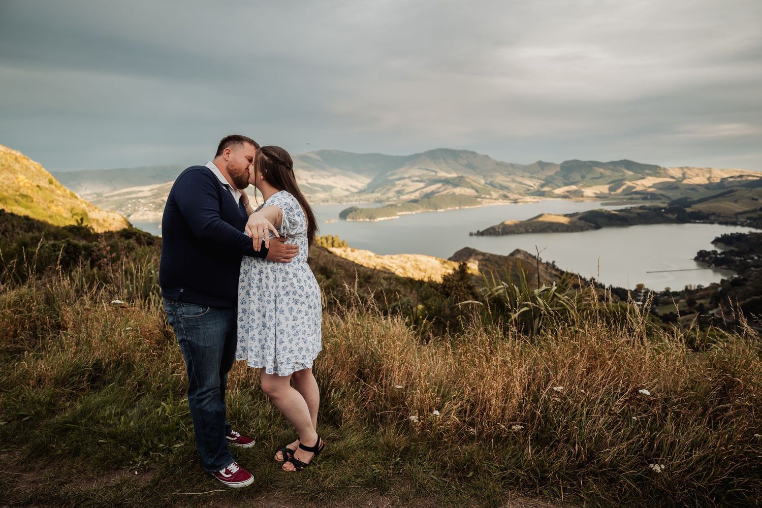 Couple kissing on a scenic hilltop with stunning coastal views and dramatic cloudy skies in the background.