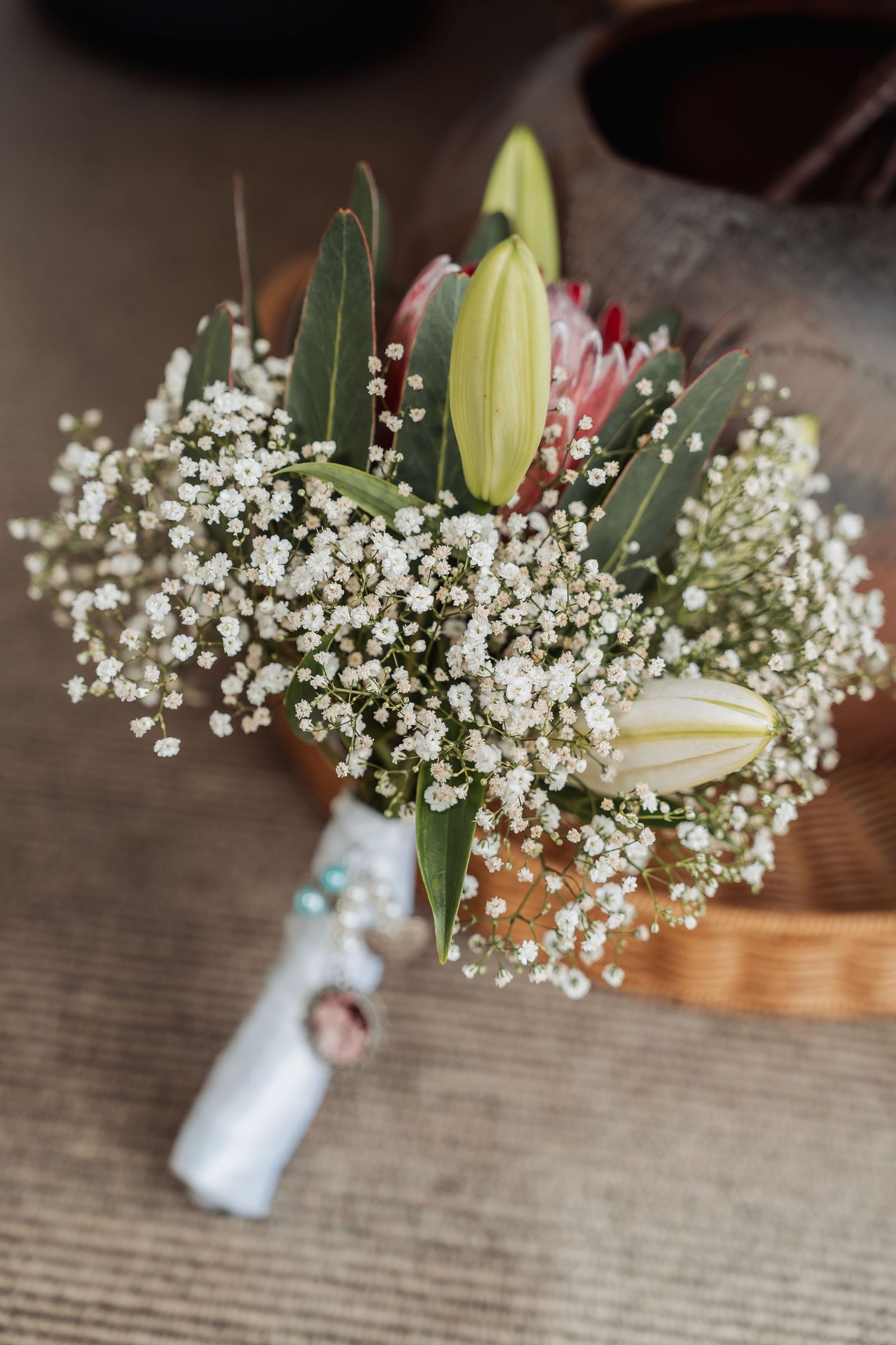 Elegant wedding boutonniere with white gypsophila, lily buds, and a pink protea wrapped in white ribbon.