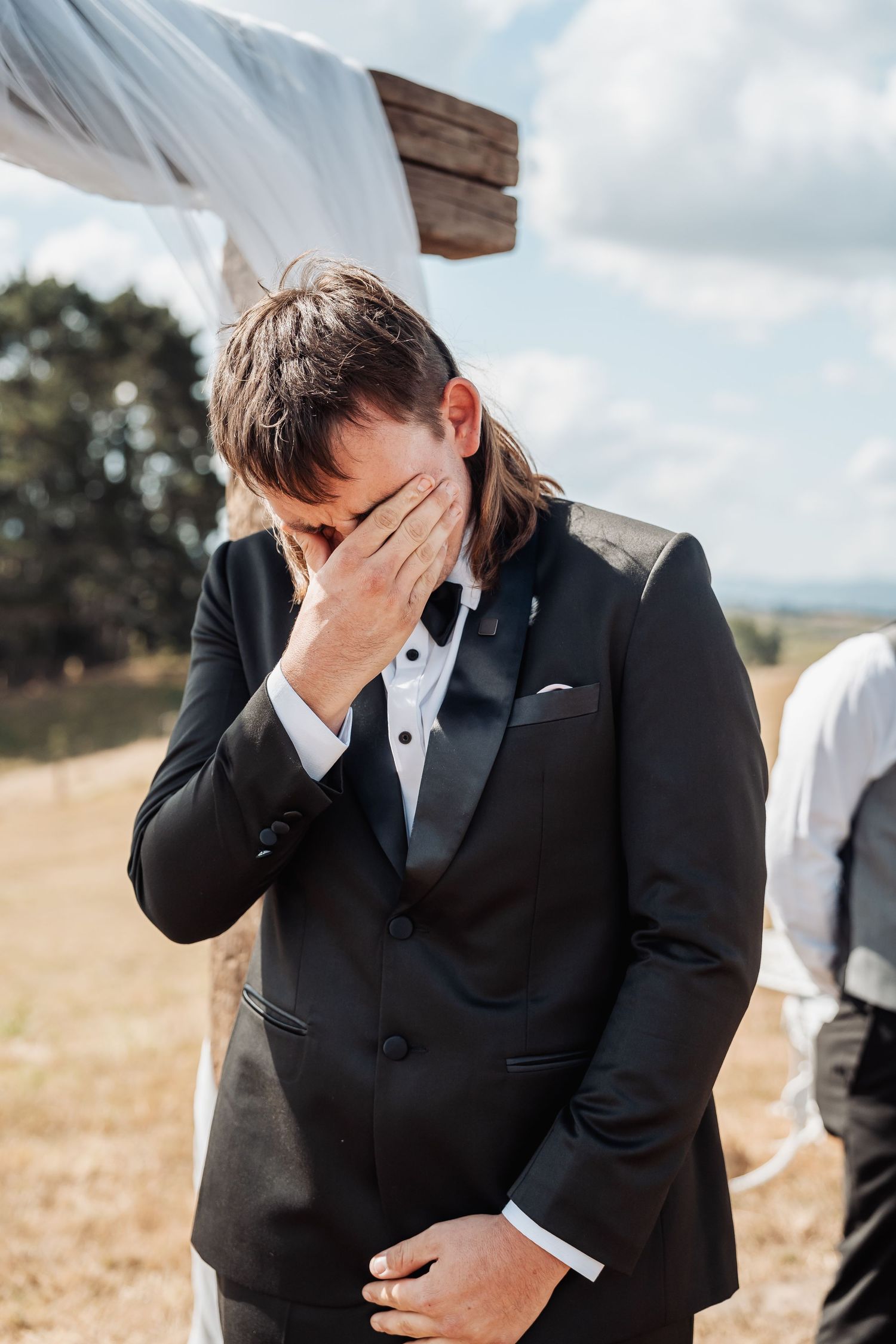 Emotional groom in black tuxedo covering face with hand at outdoor wedding ceremony.