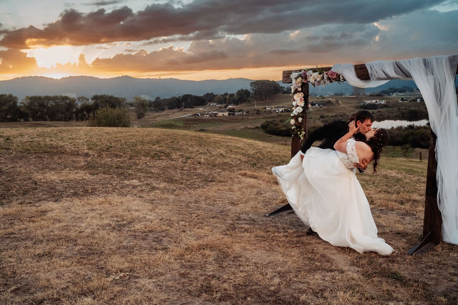 Bride and groom share romantic dip kiss under floral arch at sunset wedding.