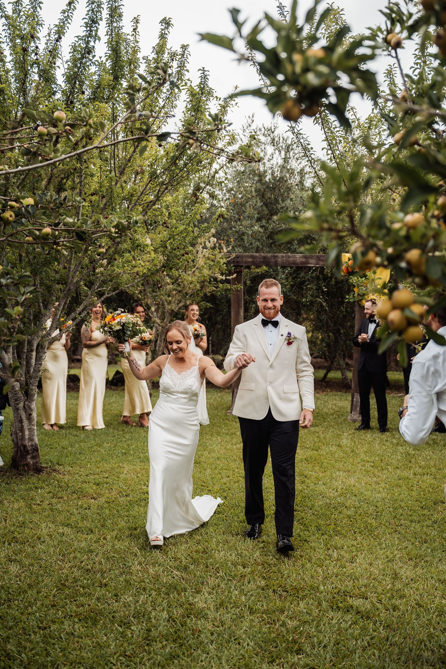Bride and groom walking hand in hand through an outdoor orchard ceremony recessional.