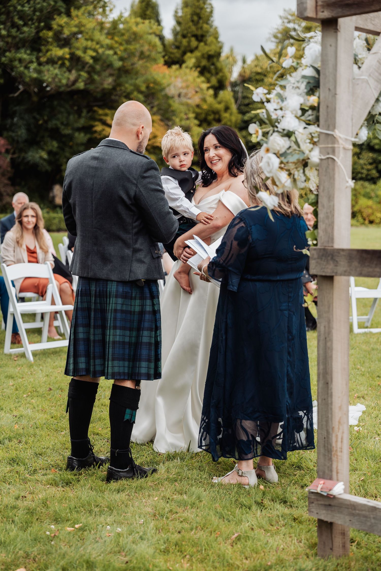 A bride and groom in a kilt exchange vows outdoors while holding a child during their garden wedding ceremony.