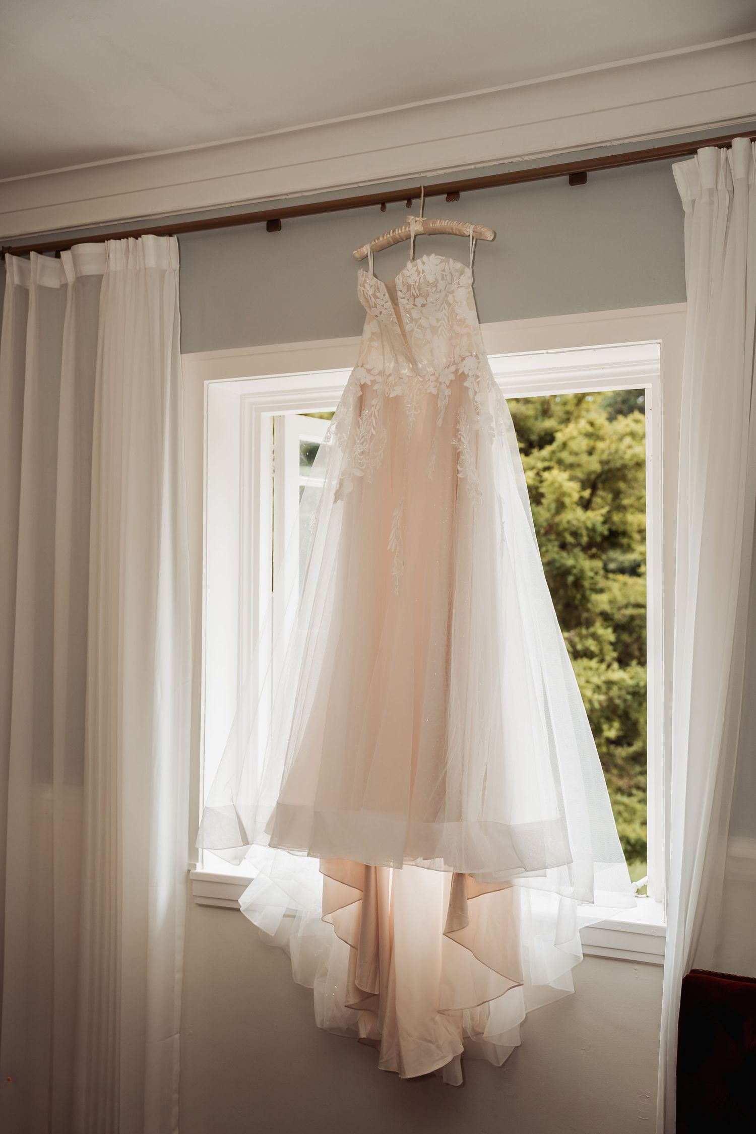 Elegant strapless lace wedding dress hanging in a sunlit window with white curtains.