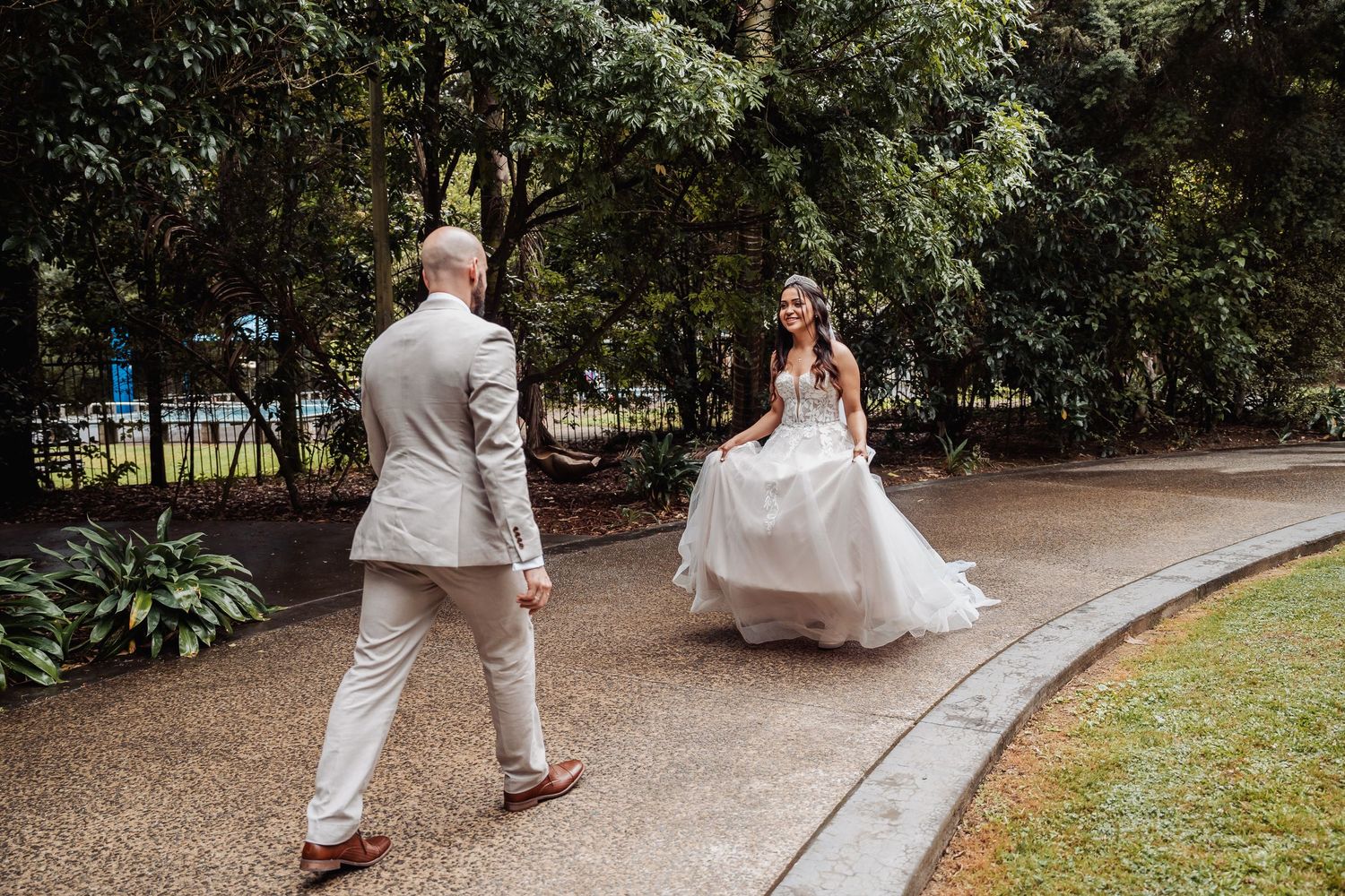 Bride in white ball gown walks toward groom on rainy garden path surrounded by lush greenery.