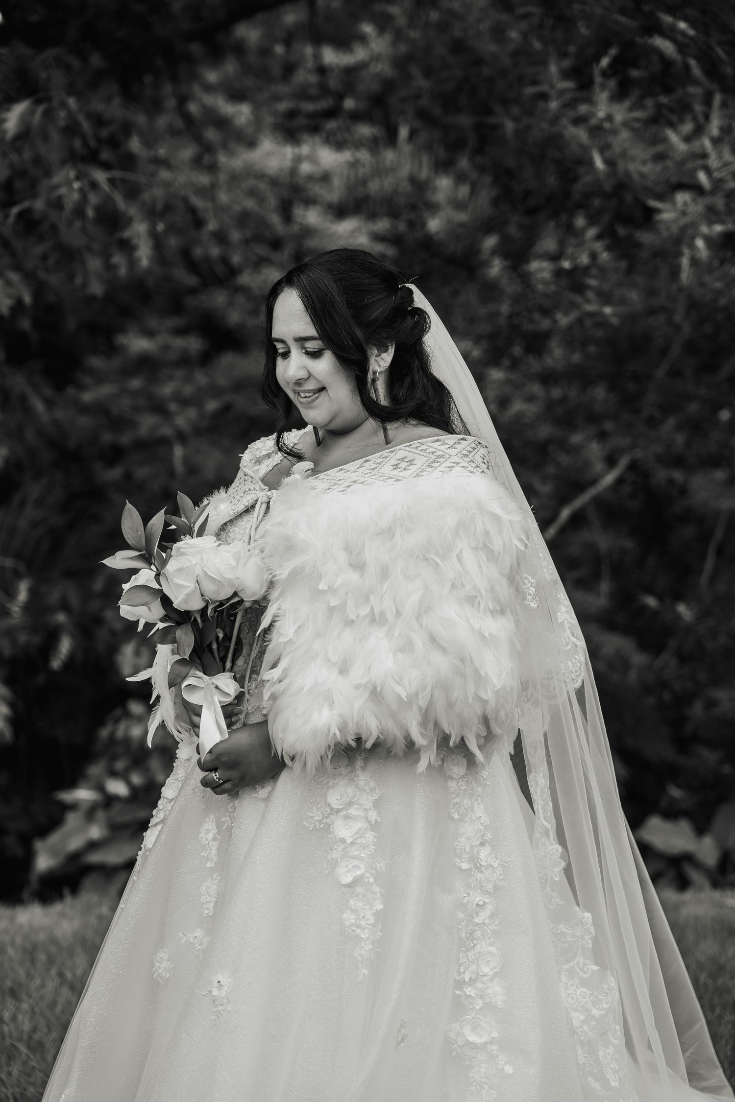 Black and white portrait of bride in feather wrap holding bouquet in garden setting.