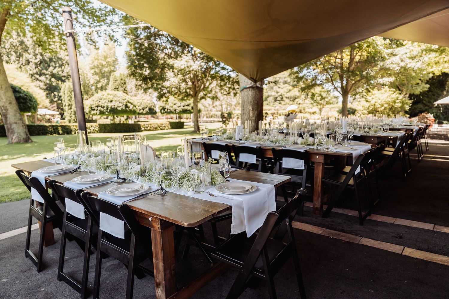 Elegant outdoor wedding reception with long wooden tables, black chairs, and glassware set under a shaded canopy.