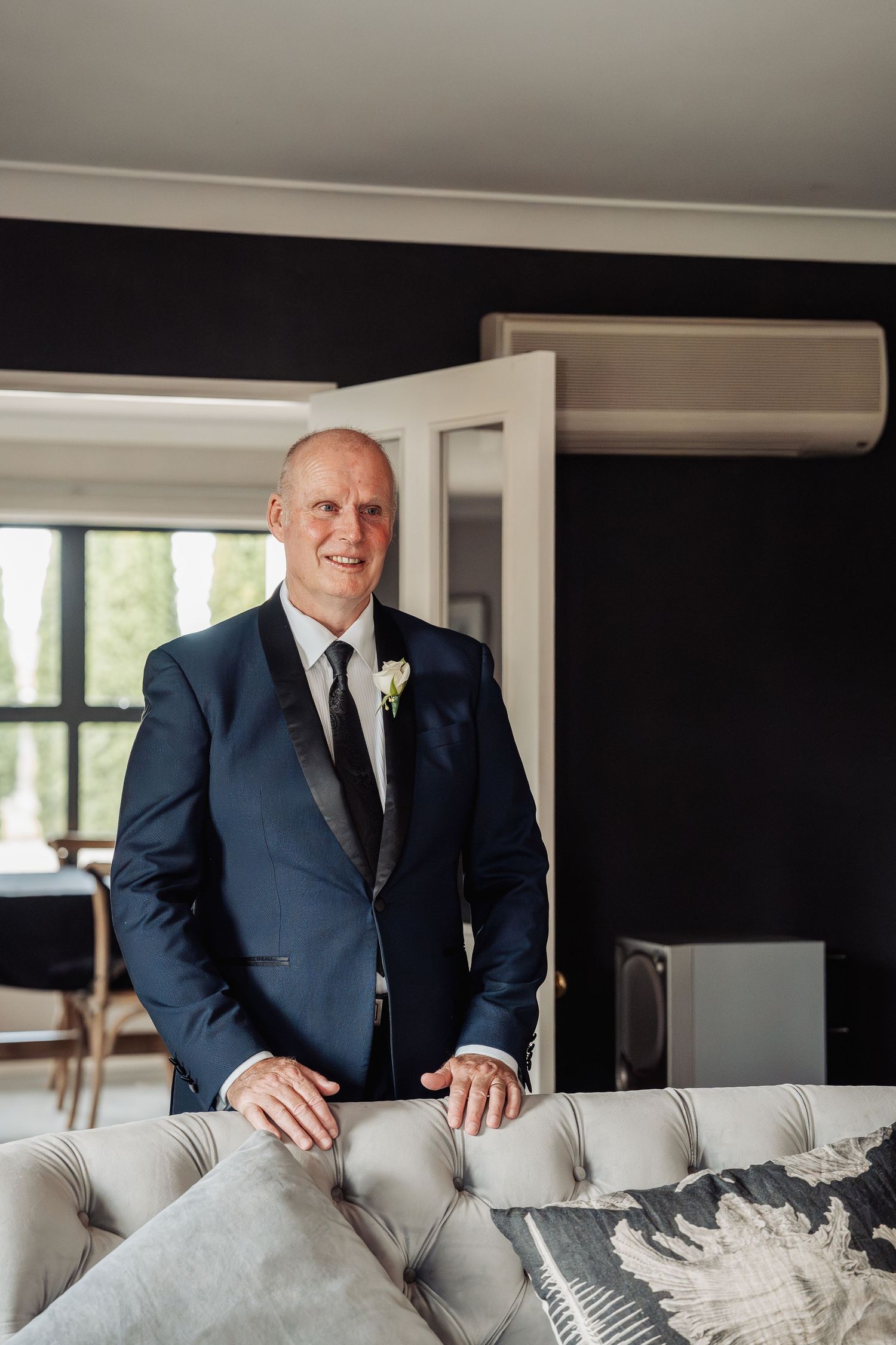 Groom in navy blue tuxedo with white boutonniere stands confidently beside a tufted sofa in elegant venue.