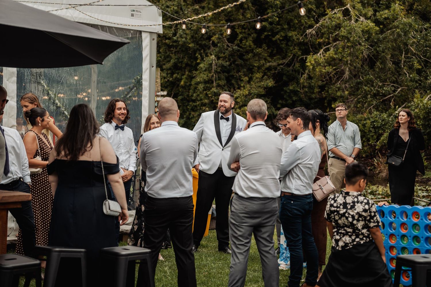 Guests mingling outdoors at a wedding reception with string lights, a tent, and lawn games in a garden setting.