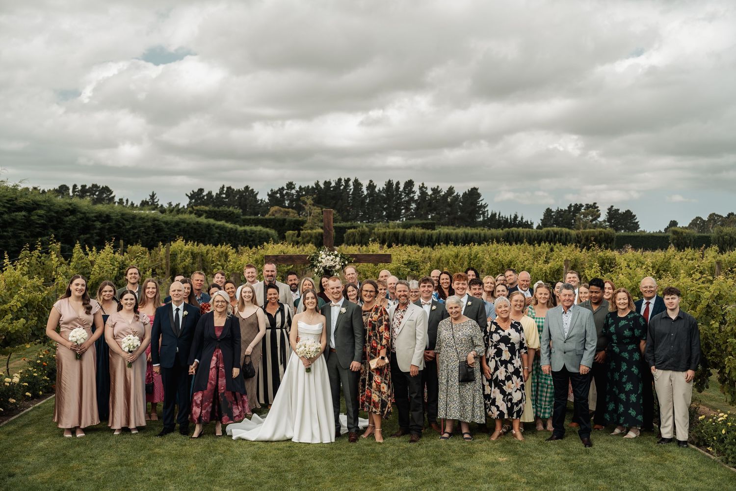 Large wedding party group photo taken outdoors with cloudy sky and lush greenery in the background.