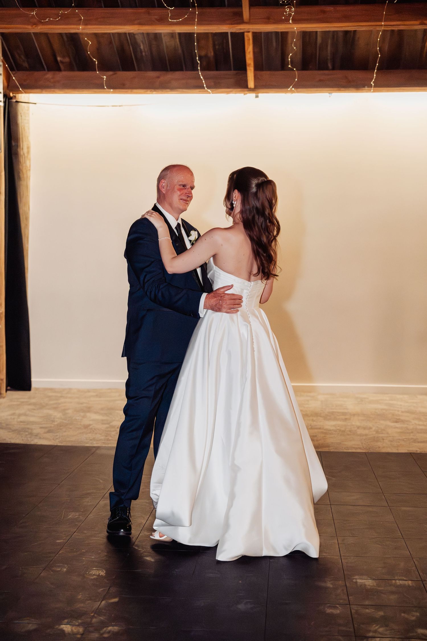 Bride and groom share their first dance, embracing closely in a warmly lit rustic venue with fairy lights overhead.