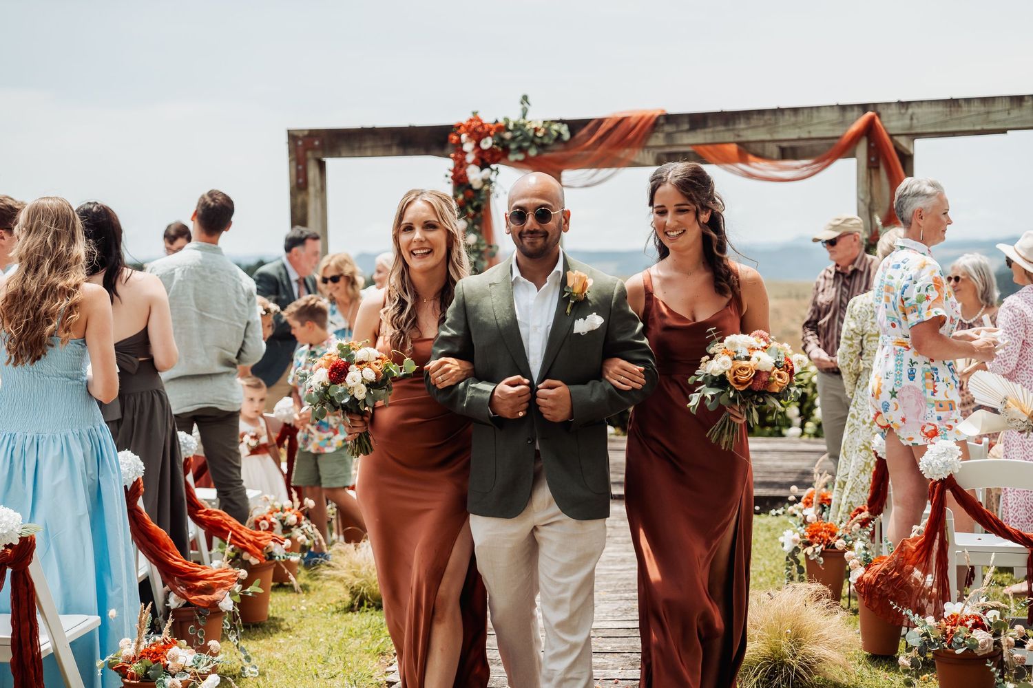 Groom walking two bridesmaids in rust dresses down the aisle at an outdoor wedding.