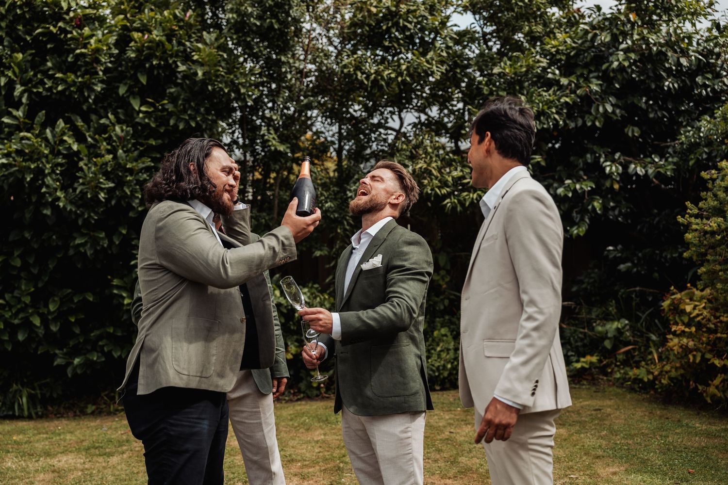 Three groomsmen celebrate outdoors as one pours champagne in a sunny garden.