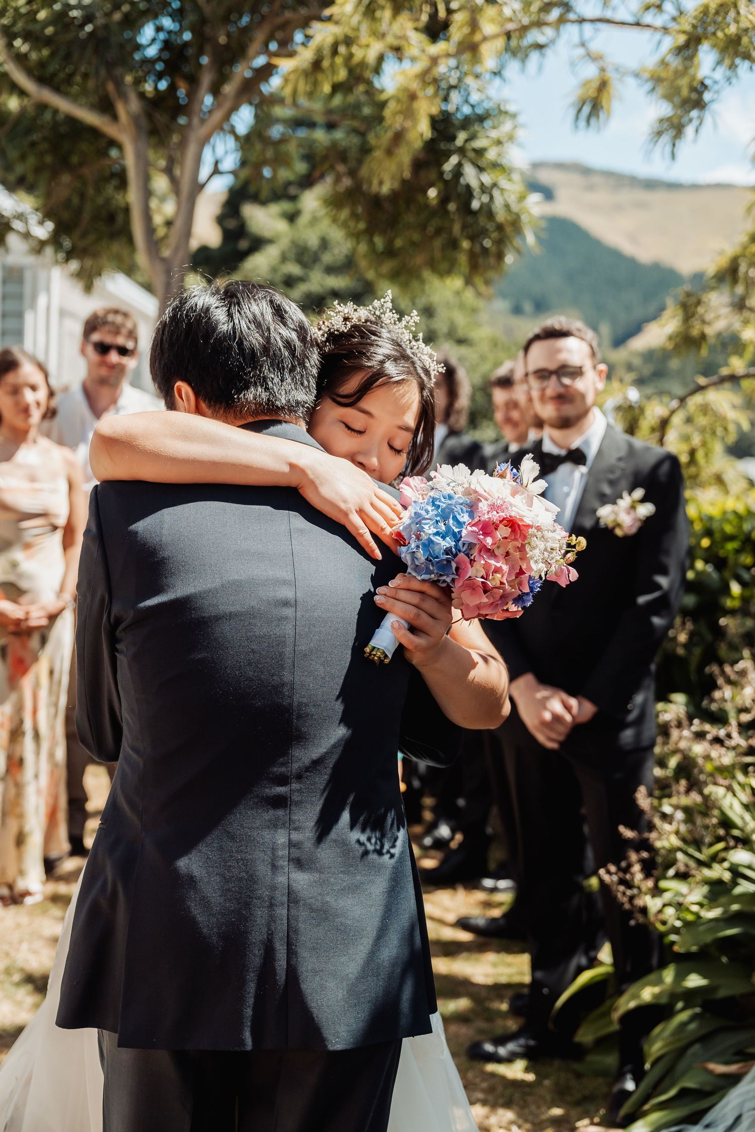 Bride hugging a guest at an outdoor wedding ceremony while holding a colorful bouquet of blue and pink flowers.