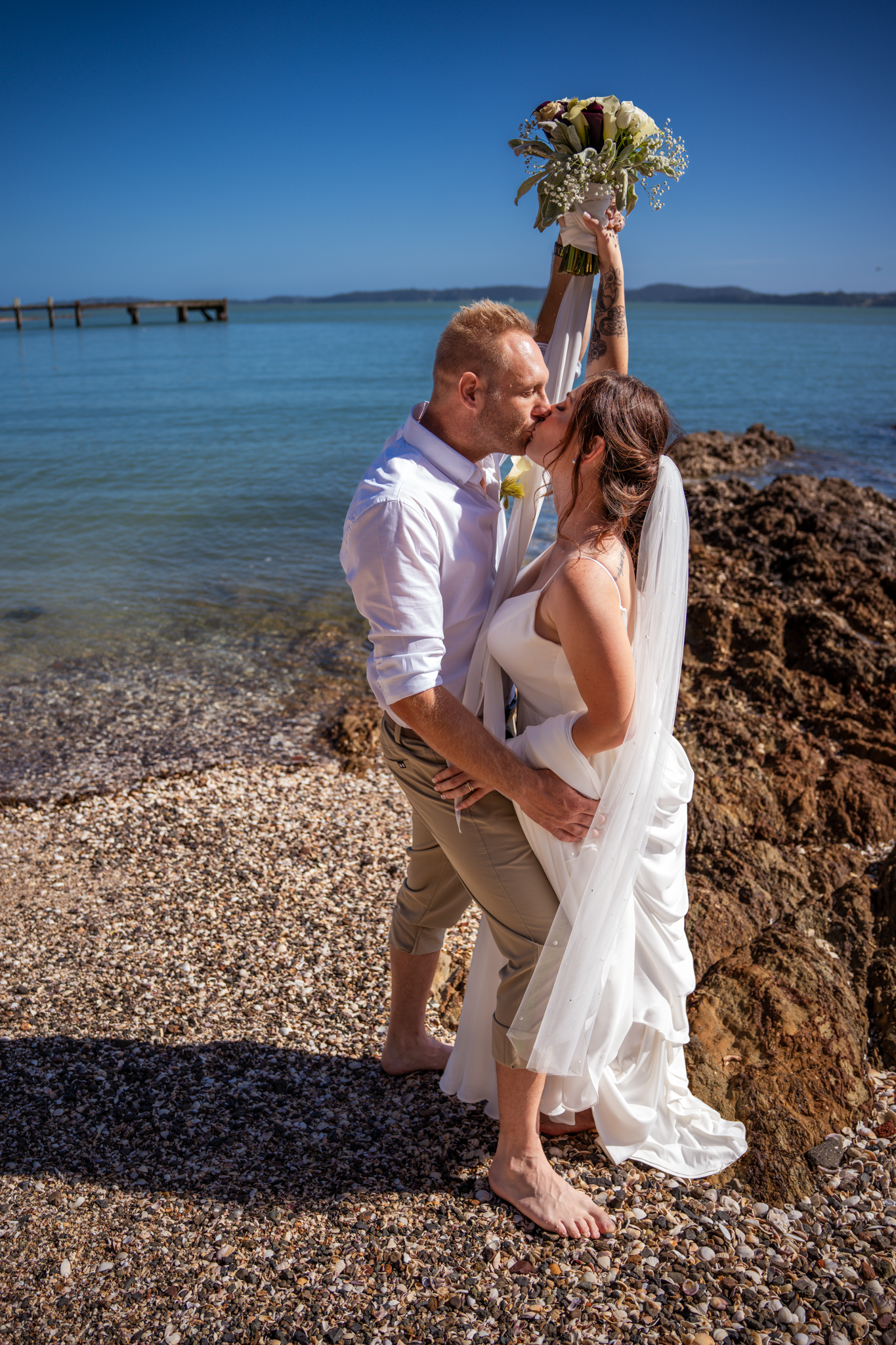 Wedding couple embraces on rocky beach shore with bouquet raised against blue ocean view.