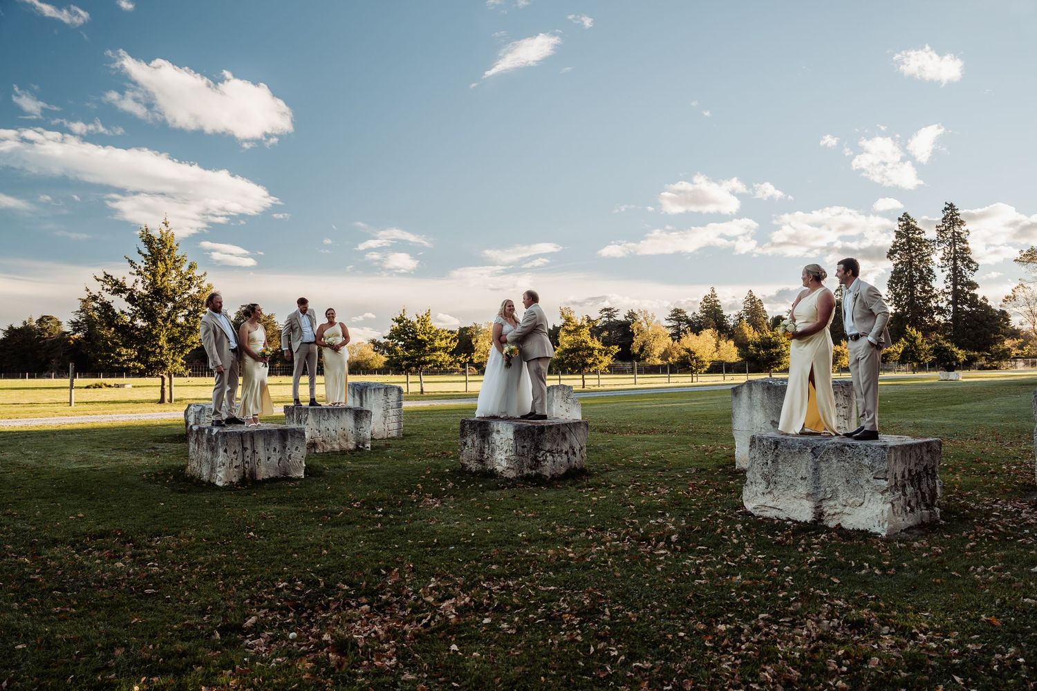 Wedding party poses on stone pedestals in open green field under blue sky at golden hour.