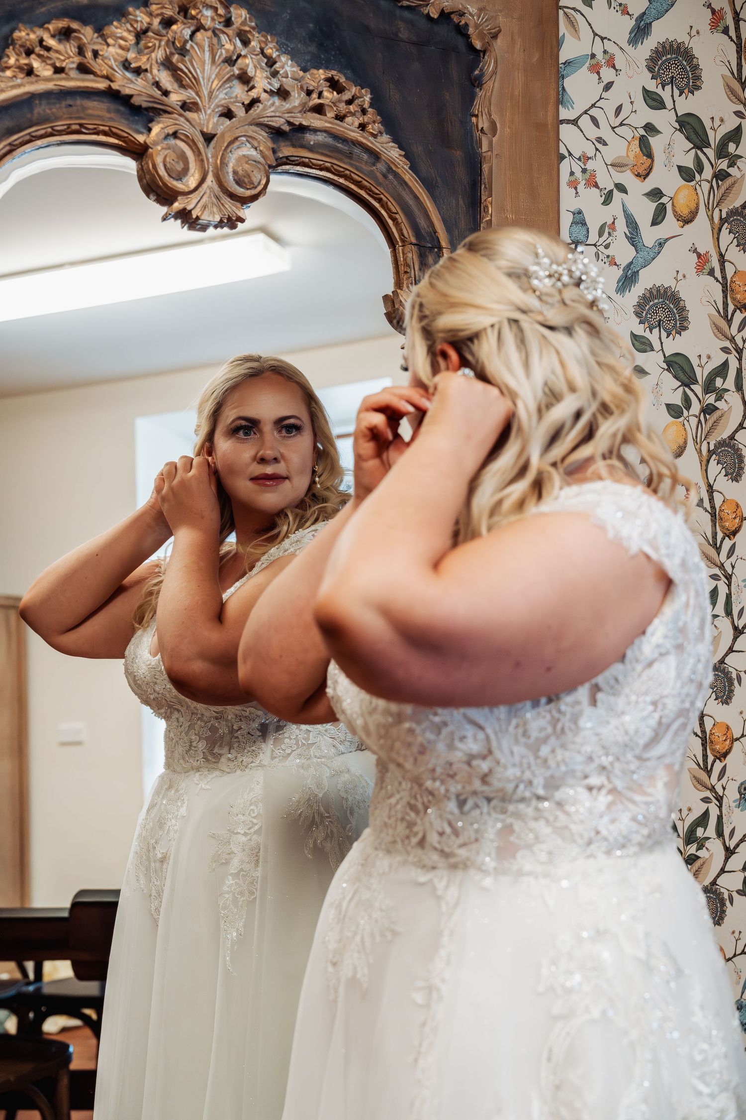Blonde bride in lace wedding dress adjusting earrings while looking in ornate gold mirror.