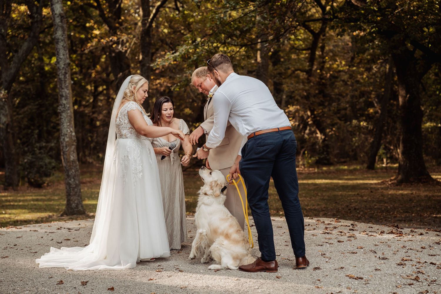 Bride and groom with officiant and golden retriever dog during outdoor wedding ceremony in autumn forest setting.