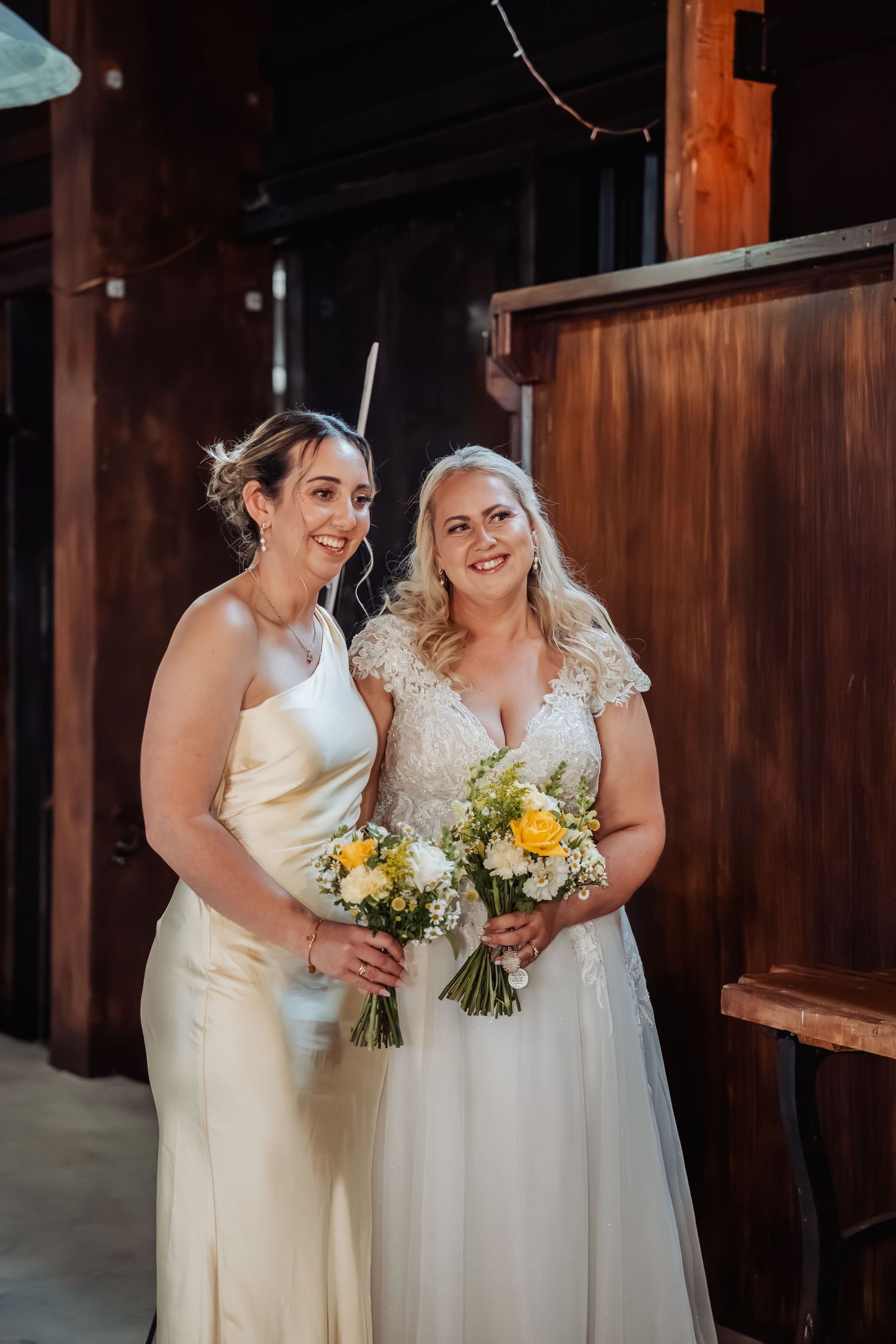 Two brides in white wedding gowns holding yellow and white bouquets pose together in a rustic barn venue.