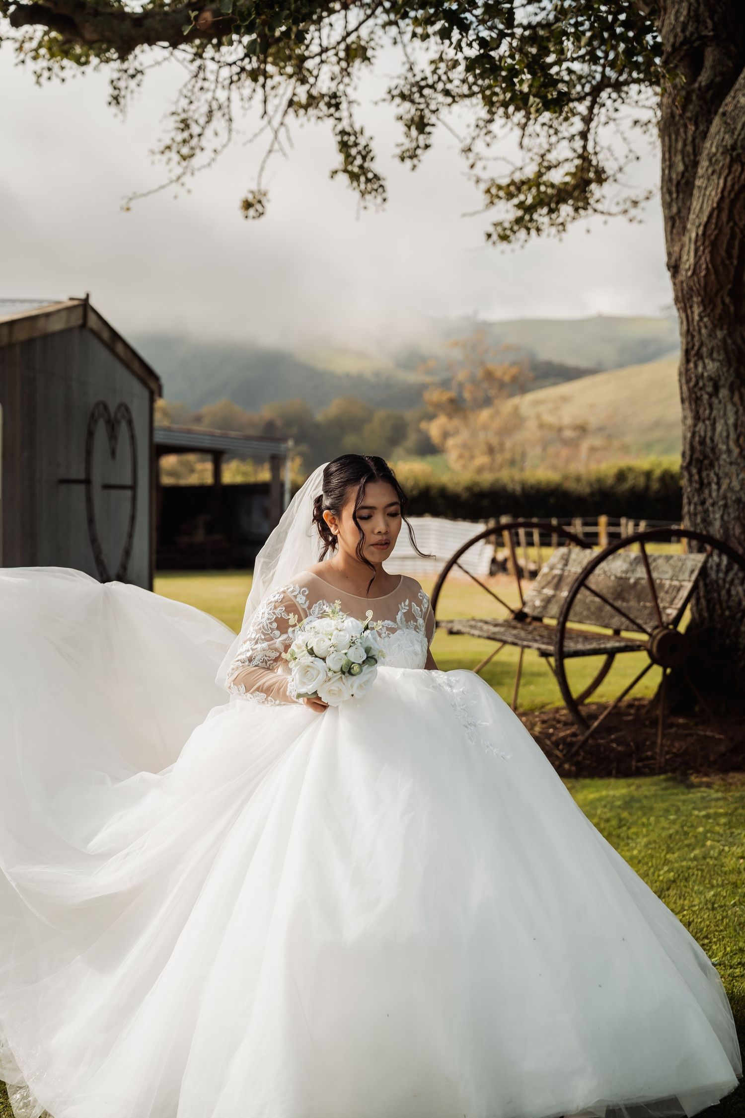 Bride in a flowing white ball gown and veil holds a bouquet outdoors with rolling hills and rustic barn backdrop.