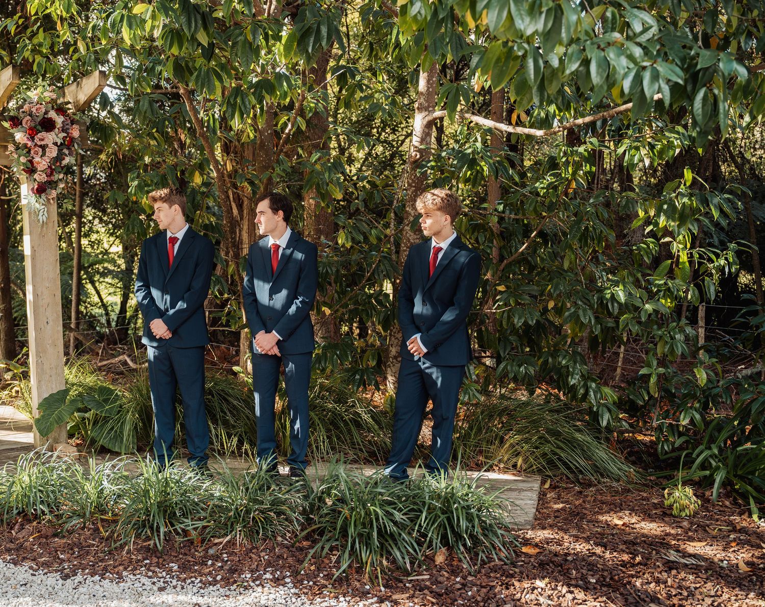 Three groomsmen in navy suits with red ties stand at outdoor wedding ceremony altar.