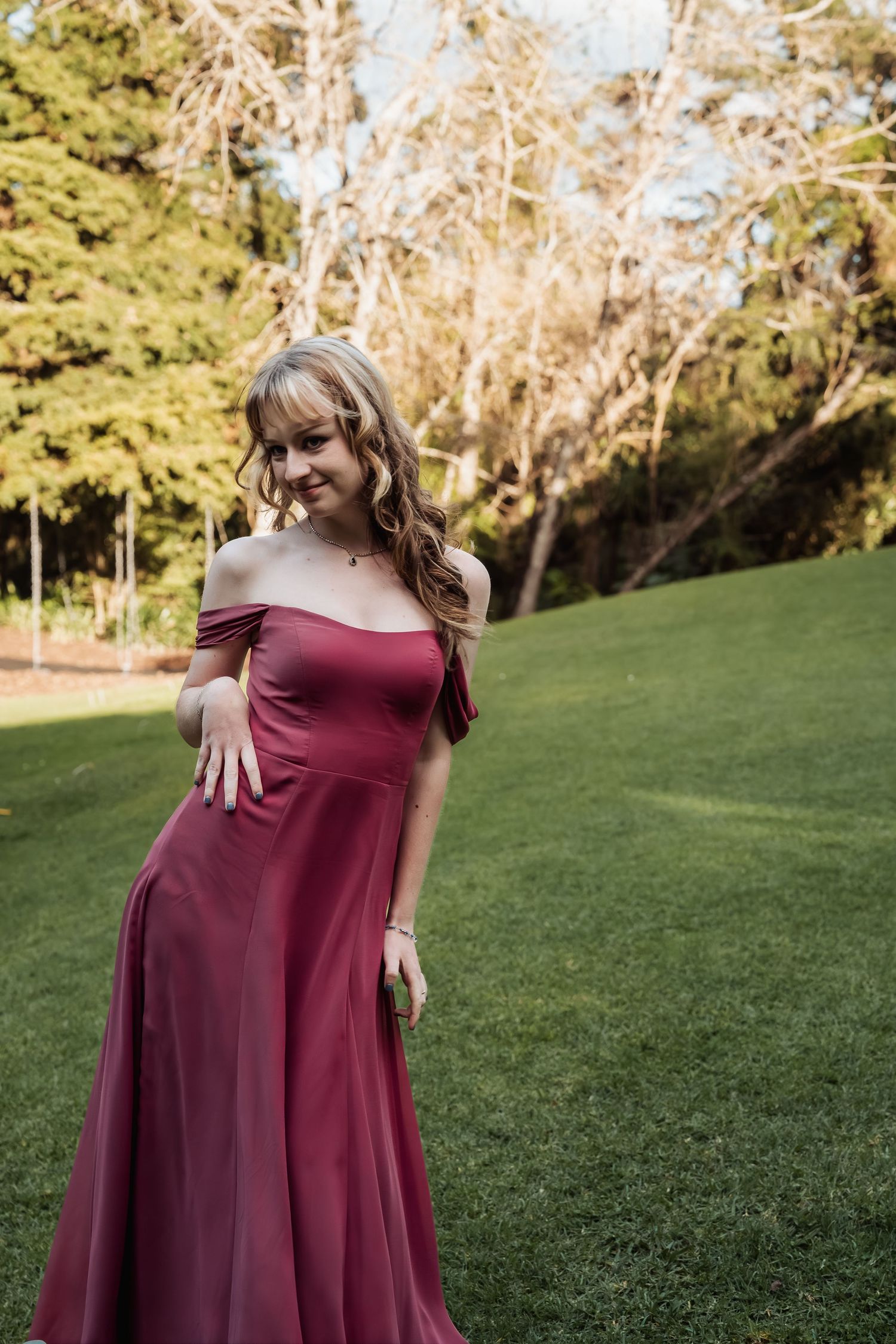 Smiling bridesmaid poses elegantly in an off-shoulder burgundy gown on a green lawn with trees in the background.