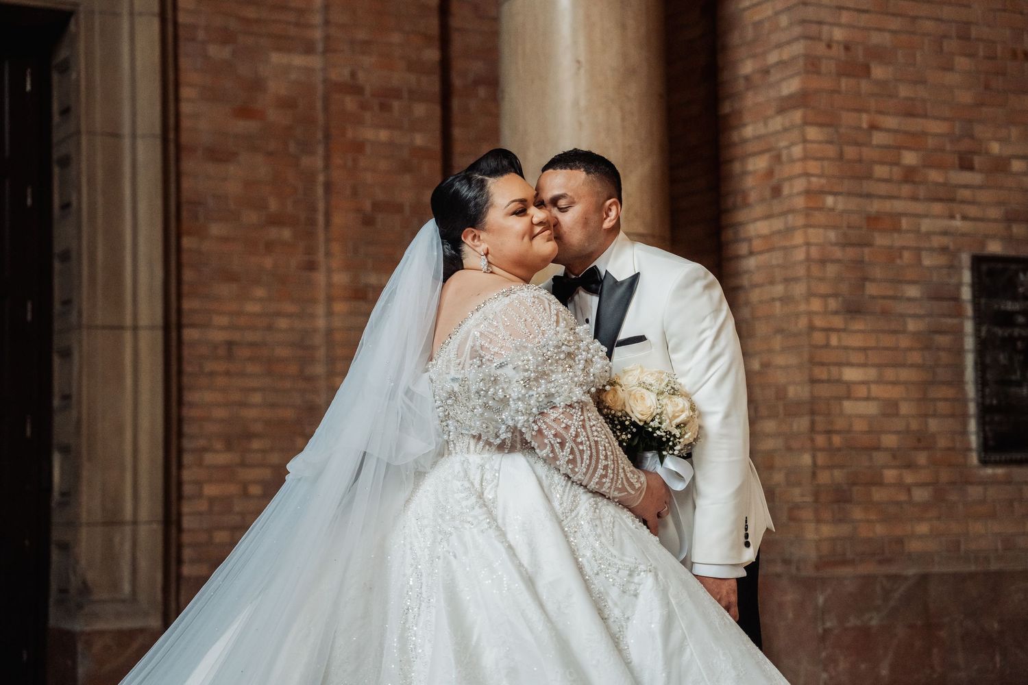Wedding couple embraces in romantic moment against rustic brick wall and columns.